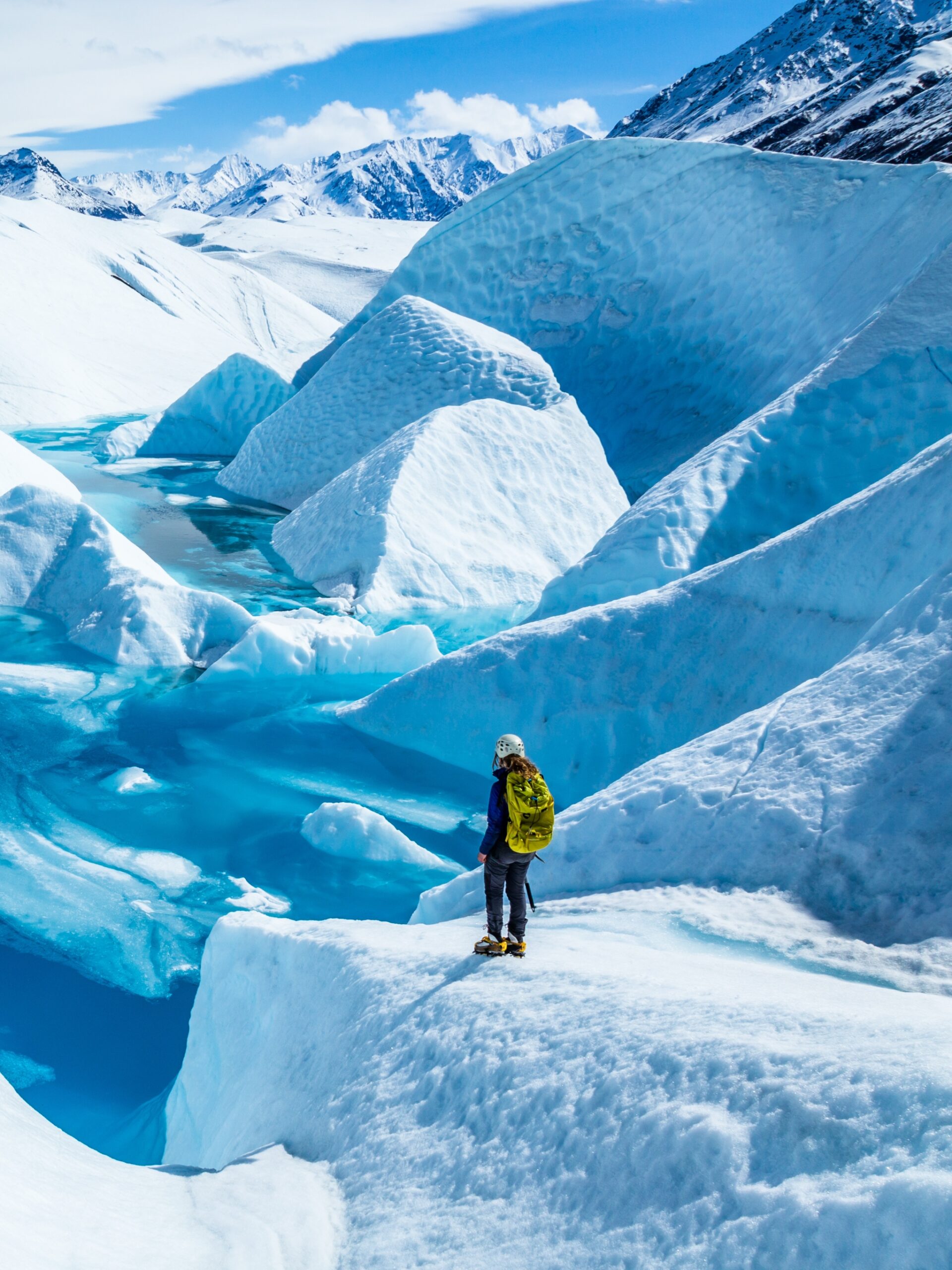 Standing near the edge of large blue pool on top of the Matanuska Glacier. A young woman holding an ice axe with a backpack and helmet looks out over the lake.